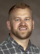 A man with short blond hair and a beard is wearing a gray checked shirt and smiling at the camera against a gray background.